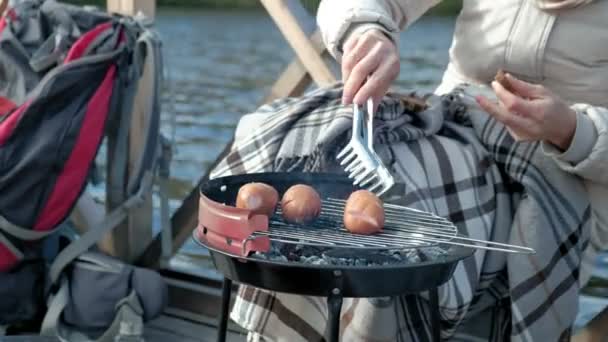 Vrouw toerist in warme kleren op de brug door de bank van de rivier met een rugzak, voorbereiding van de worstjes op de barbecue, picknick, actieve ontspanning, gezonde levensstijl. Travel concept, close-up — Stockvideo