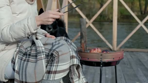 Touristin in warmer Kleidung auf der Brücke am Flussufer mit Rucksack, Würstchen auf dem Grill zubereiten, Picknick, neben einem Hund sitzen, aktive Freizeit, gesunder Lebensstil. Reisekonzept — Stockvideo