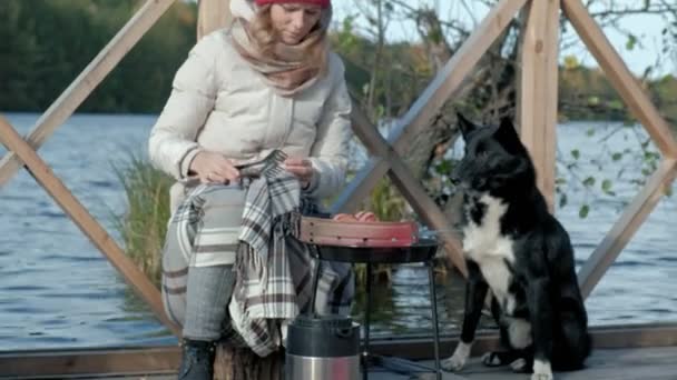 Una turista donna in abiti caldi su un ponte vicino alla riva del fiume con uno zaino, preparare salsicce alla griglia, pic-nic, nutre il cane, riposo attivo, stile di vita sano. Concetto di viaggio — Video Stock