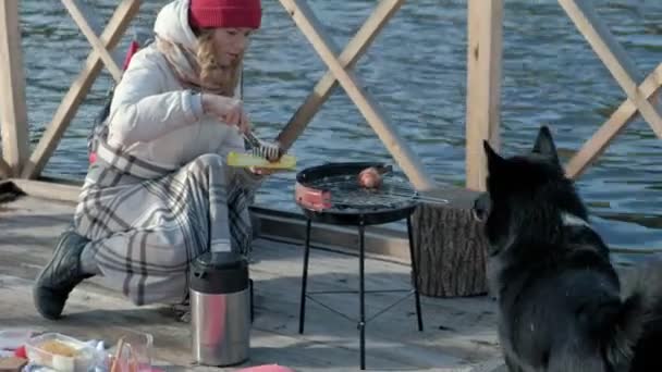 Femme touriste en vêtements chauds sur le pont au bord de la rivière avec un sac à dos, préparer des saucisses sur le gril, pique-nique, assis à côté d'un chien, loisirs actifs, mode de vie sain. Concept de voyage — Video