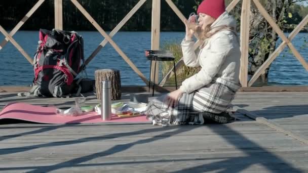 Woman tourist in warm clothes on the bridge by the river bank with a backpack, drinking hot tea, picnic, active rest, healthy lifestyle. Travel concept, close-up — Stock Video