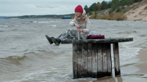Mujer joven con ropa de abrigo sentada junto al océano, en una bobina de madera, bebiendo té caliente de un termo, clima frío, tormenta — Vídeos de Stock