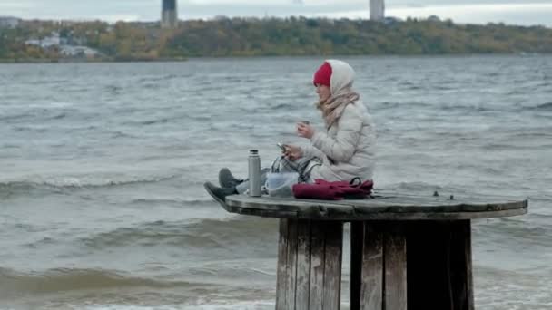 Jonge vrouw in warme kleren zitten op de Oceaan kust, op een houten spoel, drinken hete thee uit een thermos, met behulp van een telefoon, koud weer, een storm — Stockvideo