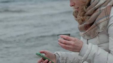 kullanarak bir telefon, soğuk hava, fırtına, yakın çekim, portre bir termos sıcak çay içme sıcak giysiler okyanusta ahşap bir rulo başına oturmuş genç kadında