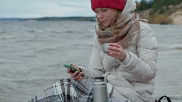 Jeune femme en vêtements chauds assise au bord de l'océan, sur une bobine de bois, buvant du thé chaud dans un thermos, en utilisant un téléphone, temps froid, tempête, gros plan, portrait — Video