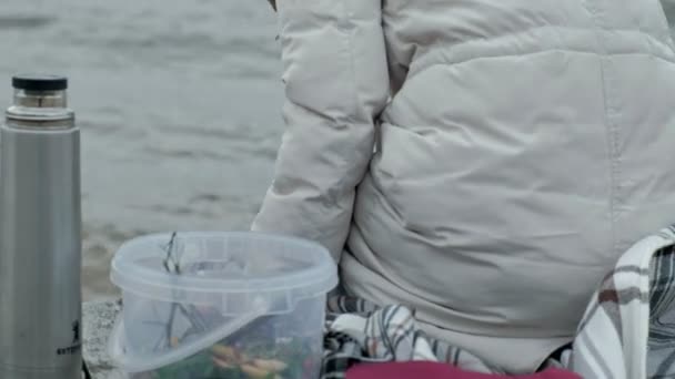Mujer joven con ropa de abrigo sentada en la orilla del océano, en una bobina de madera, bebiendo té caliente de un termo, tocando el agua con su pie, clima frío, tormenta — Vídeos de Stock
