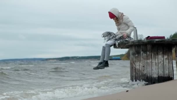 Jeune femme en vêtements chauds assise sur le bord de l'océan, sur une bobine de bois, buvant du thé chaud d'un thermos, touchant l'eau avec son pied, temps froid, tempête — Video