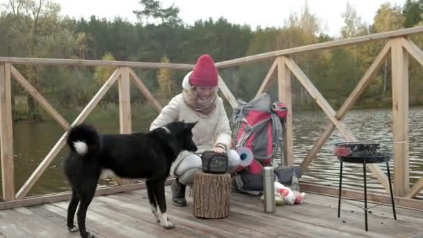 Eine Touristin auf der Brücke am Flussufer streichelt einen Hund, kocht Essen, holt Sachen aus dem Rucksack, Picknick, Outdoor-Aktivitäten und einen gesunden Lebensstil. Reisekonzept — Stockvideo