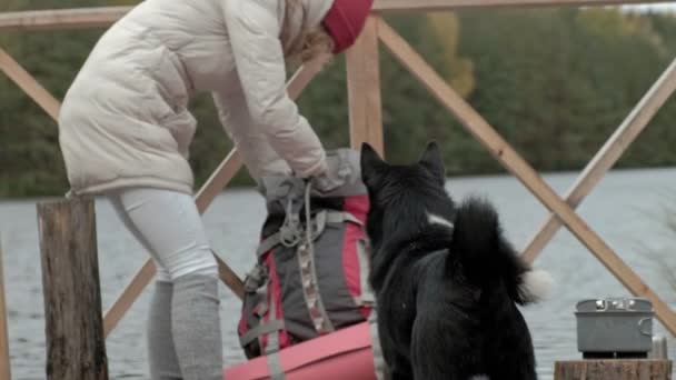 Uma turista se senta na ponte perto da margem do rio, perto de um cão, prepara comida, tira coisas de sua mochila, piquenique, atividades ao ar livre e um conceito de viagem de estilo de vida saudável, close-up — Vídeo de Stock