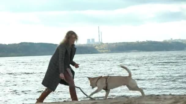 Ung kvinna i en kappa med en tjej med lockigt hår, Mamma och dotter, springa, leka med en brun hund på stranden, grubbla sin pinne, kallt väder — Stockvideo