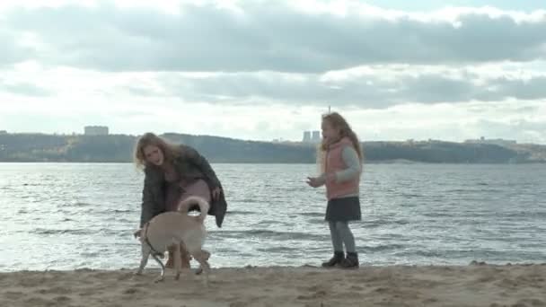 Giovane donna in un cappotto con una ragazza con i capelli ricci, mamma e figlia, correre, giocare con un cane marrone sulla spiaggia, covare il suo bastone, tempo freddo — Video Stock