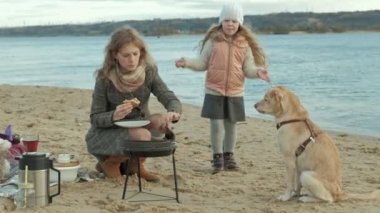 Anne ve kızı, kıvırcık saçlı bir kız ırmak, okyanus, piknik yaptı sahilde bir ceket genç bir kadın oturur, yakın, soğuk hava doğa, aç bir köpek Hamburger yiyor yürüyor