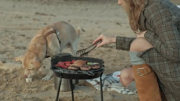 Młoda kobieta w płaszcz i dziewczyna z kręconymi włosami, matka z córką, siedząc na plaży przez rzeki, ocean, miał piknik, gotowanie mięsa i warzywa na grillu, dziewczyna gra z psem — Wideo stockowe