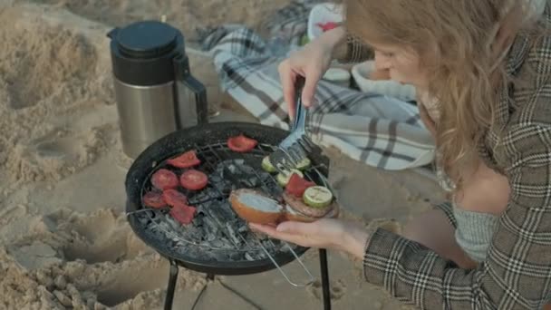 Молода жінка в пальто сидить на пляжі біля річки, океан, має пікнік, готує м'ясо і овочі на грилі, збирає бургер в природі, холодну погоду — стокове відео