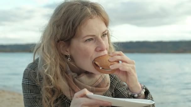 Mujer joven en un abrigo se sienta en la playa junto al río, el océano, tiene un picnic, come una hamburguesa en la naturaleza, tiempo frío — Vídeo de stock