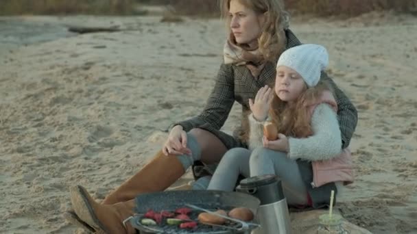 Jonge vrouw met een vacht met een meisje met krullend haar, moeder met dochter, op het strand aan de rivier, Oceaan, had een picknick, rust, weekend, koud weer — Stockvideo