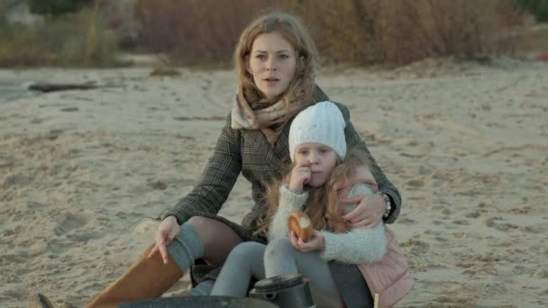Genç kadın bir ceket anne ile kızı, kıvırcık saçlı bir kızla sahilde nehrin yanında, okyanus, vardı bir piknik, dinlenme, hafta sonu, soğuk hava — Stok video