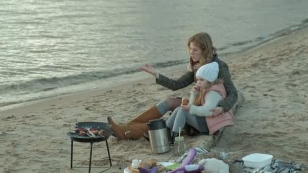 Junge Frau im Mantel mit einem Mädchen mit lockigem Haar, Mutter mit Tochter, am Strand am Fluss, Meer, Picknick, Erholung, Wochenende, kaltes Wetter — Stockvideo