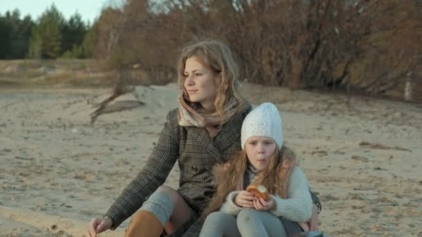 Jonge vrouw met een vacht met een meisje met krullend haar, moeder met dochter, op het strand aan de rivier, Oceaan, had een picknick, rust, weekend, koud weer — Stockvideo