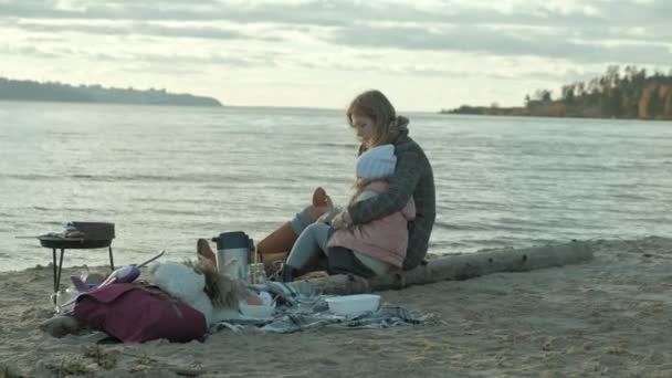 Jonge vrouw met een vacht met een meisje met krullend haar, moeder met dochter, op het strand aan de rivier, Oceaan, had een picknick, rust, weekend, koud weer — Stockvideo