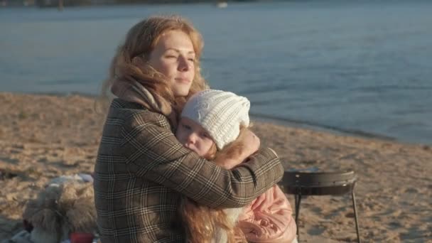 Молодая женщина в пальто с девушкой с вьющимися волосами, мать с дочерью, на пляже у реки, океан, устроила пикник, отдых, выходные, холодная погода — стоковое видео