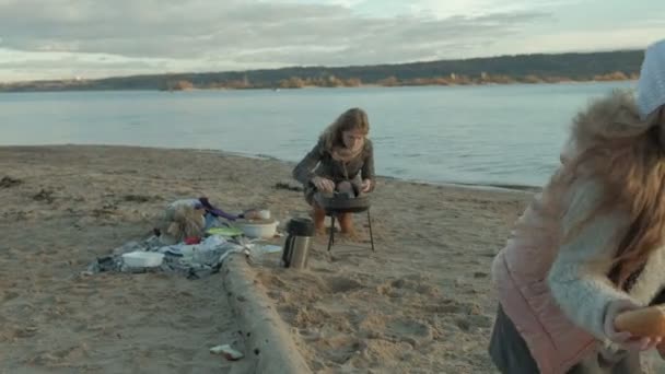 Giovane donna in cappotto con una ragazza con i capelli ricci, madre con figlia, sulla spiaggia vicino al fiume, oceano, ha fatto un picnic, riposo, fine settimana, tempo freddo — Video Stock