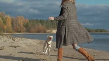 Genç kadın bir paltoyla kıvırcık saçlı bir kız, anne ve kızı, koş, kahverengi bir köpek sahilde oynamak, köpek bir sopa su, soğuk hava dışarı çekmek