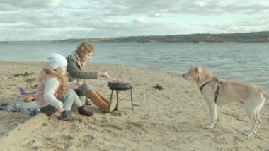 Genç kadın bir ceket ve kıvırcık saçlı, Anne kızı, olan bir kızla oturup ırmak kenarına, sahilde okyanus, ızgara et pişirmek, piknik, kız beslemek köpek, soğuk hava