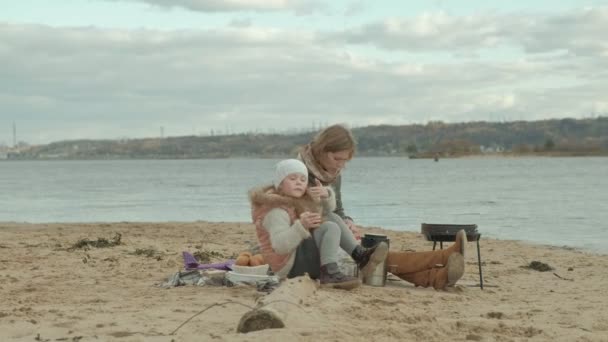 Νεαρή γυναίκα με ένα παλτό με μια κοπέλα με τα σγουρά μαλλιά, μαμά και κόρη, κάθονται στην παραλία δίπλα στον ποταμό ωκεανό, έχοντας ένα πικ-νικ, πίνοντας ζεστό τσάι, κοπής λαχανικών, κρύο — Αρχείο Βίντεο