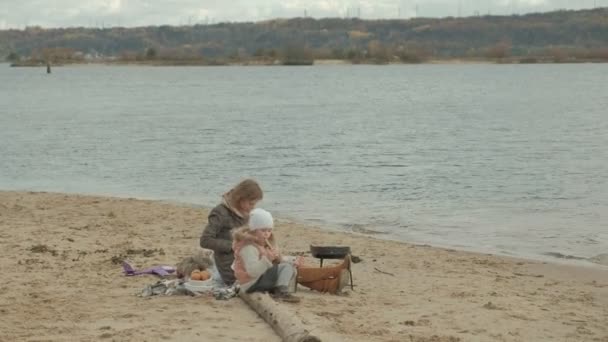 Mujer joven en un abrigo con una chica con el pelo rizado, mamá y su hija, sentado en la playa junto al río, el océano, teniendo un picnic, beber té caliente, cortar verduras, clima frío — Vídeos de Stock