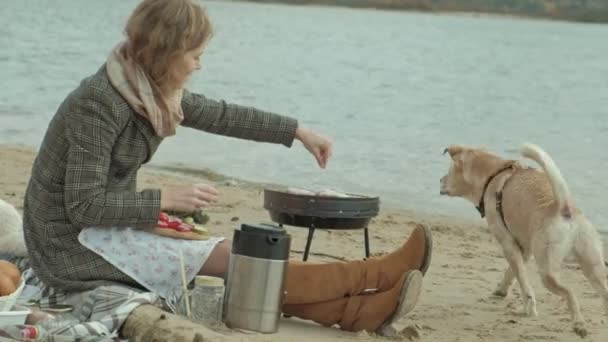 Молода жінка в пальто сидить на пляжі біля річки, в океані, має пікнік, готує овочі та м'ясо на грилі, собака грає поблизу, холодна погода — стокове відео
