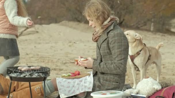 母亲与女儿, 一个年轻的妇女在大衣坐在海滩上的河流, 海洋, 做了野餐, 厨师蔬菜和肉类在烧烤, 旁边的女孩卷曲的头发玩狗, 冷 — 图库视频影像
