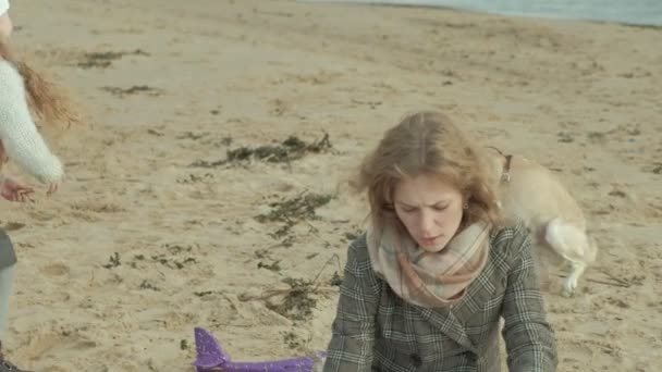 Young woman in a coat with a girl with curly hair, mom and daughter, sitting on the beach by the river, ocean, having a picnic, drinking hot tea, cutting vegetables, near a dog, cold weather — 图库视频影像