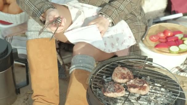 Ung kvinna i jacka ligger på stranden av floden, ocean, har picknick, kockar kött på grillen, kallt väder — Stockvideo