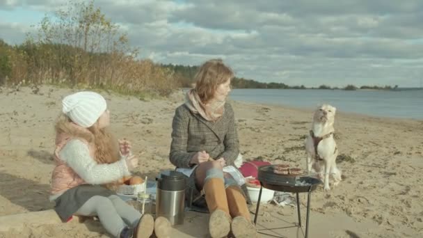 Junge Frau im Mantel und ein Mädchen mit lockigem Haar, Mutter mit Tochter, sitzt am Strand am Fluss, Meer, hatte ein Picknick, Fleisch kochen auf dem Grill, neben einem Hund sitzen, kaltes Wetter — Stockvideo