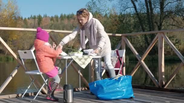 Una mujer joven y una niña en ropa de abrigo, una madre y una hija, tuvieron un picnic en la orilla del río en un puente de madera, la colocación de platos, fines de semana, clima frío, recreación al aire libre — Vídeo de stock