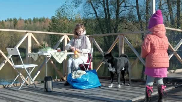 Una mujer joven y una niña en ropa de abrigo, madre e hija, tuvieron un picnic en la orilla del río en un puente de madera, preparar la comida, la niña alimenta al perro, fines de semana, clima frío, al aire libre — Vídeos de Stock