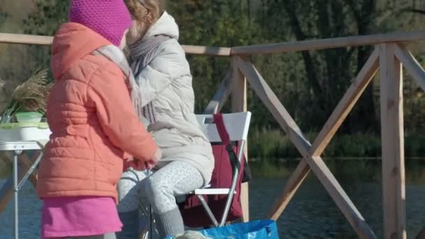 Молода жінка в теплому одязі, мати і дочка, провели пікнік на березі річки на дерев'яному мосту, приготували їжу, дівчина годує собаку, вихідні, холодну погоду, відпочинок на відкритому повітрі, туризм — стокове відео