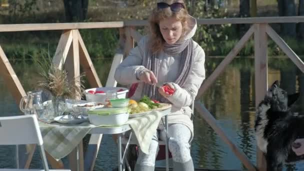 Молодая женщина в теплой одежде, пикник на берегу реки на деревянном мосту, резка овощей, собака играет поблизости, выходные, холодная погода, кемпинг, туризм — стоковое видео