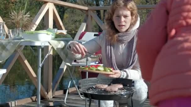 Młoda kobieta w ciepłe ubrania gotowania warzywa z grilla, chodzenie przez psa, piknik nad rzeką na drewniany most, weekend, zimnie, Kemping, turystyka — Wideo stockowe