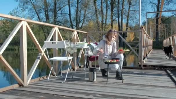 Young woman in warm clothes, cooking vegetables and meat on the grill, next to a girl playing with a dog, mother and daughter, picnic on the river bank on a wooden bridge, weekend, cold weather — Stock Video