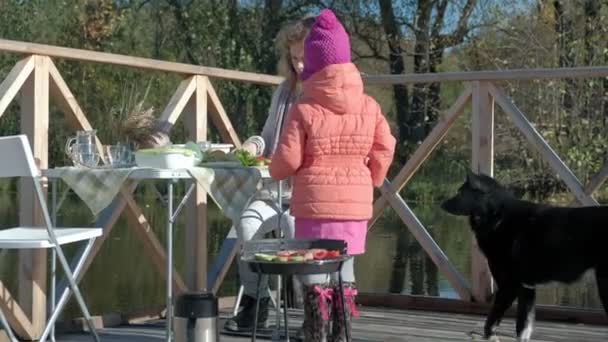 Eine junge Frau und ein kleines Mädchen in warmer Kleidung, Gemüse und Fleisch auf dem Grill kochend, ein Hund, der in der Nähe spielt, eine Mutter und eine Tochter, ein Picknick am Fluss auf einer Holzbrücke, ein Wochenende, kalt — Stockvideo