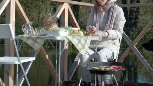 Uma jovem e uma menina em roupas quentes, cozinhar legumes e carne na grelha, um cão brincando nas proximidades, uma mãe e uma filha, um piquenique junto ao rio em uma ponte de madeira, um fim de semana, frio — Vídeo de Stock