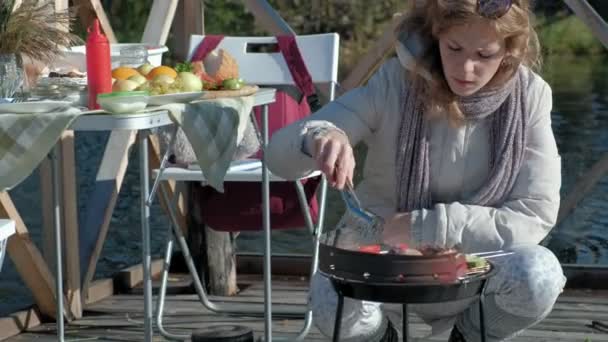 穿着暖和衣服的年轻女子, 在烤架上准备蔬菜和肉, 附近遛狗, 在一座木桥上的河岸野餐, 周末, 寒冷的天气, 户外娱乐, 旅游 — 图库视频影像