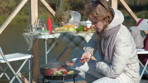 Mujer joven en ropa de abrigo, prepara verduras y carne en la parrilla, un perro pasea cerca, un picnic en la orilla del río en un puente de madera, un fin de semana, tiempo frío, recreación al aire libre, turismo — Vídeos de Stock