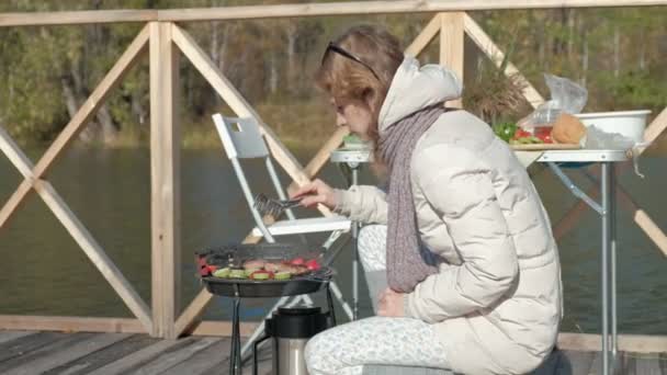 Молода жінка в теплому одязі, готує овочі та м'ясо на грилі, пікнік на березі річки на дерев'яному мосту, вихідні, холодна погода, відпочинок на відкритому повітрі, туризм — стокове відео