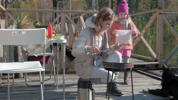 母女俩 一个年轻的女人和一个穿着暖和衣服的小女孩 在烤架上准备蔬菜和肉 用电话 在附近遛狗 在河边的木桥上野餐 — 图库视频影像