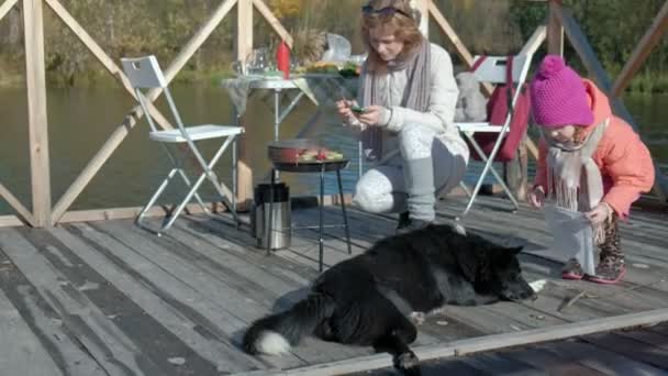 Madre e hija, una joven y una niña en ropa de abrigo, preparando verduras y carne en la parrilla, usando el teléfono, paseando a un perro cerca, un picnic junto al río en un puente de madera — Vídeos de Stock