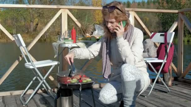 Молодая женщина в теплой одежде, готовит овощи и мясо на гриле, пользуется телефоном, пикник на берегу реки на деревянном мосту, выходные, холодная погода, кемпинг, туризм — стоковое видео
