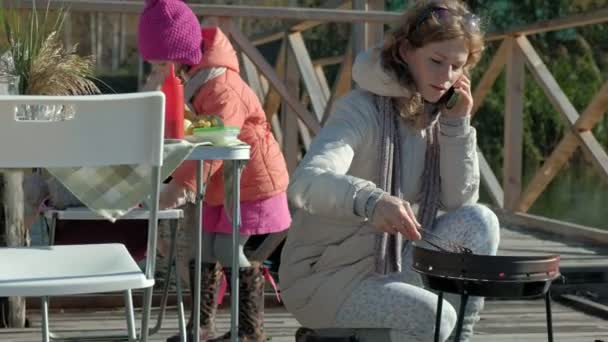 Moeder en dochter, een jonge vrouw en een klein meisje in warme kleren, voorbereiding van groenten en vlees op de grill, met behulp van de telefoon, het lopen van een hond in de buurt, een picknick aan de rivier op een houten brug — Stockvideo